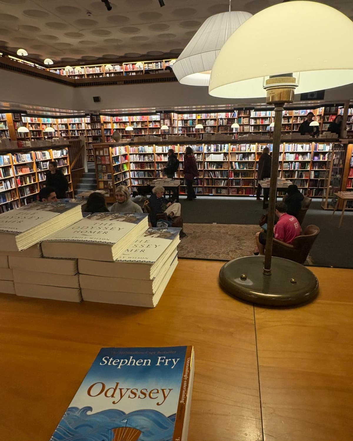 A bookshop reading room, readers in armchairs under lamps with a table of stock in the foreground.
