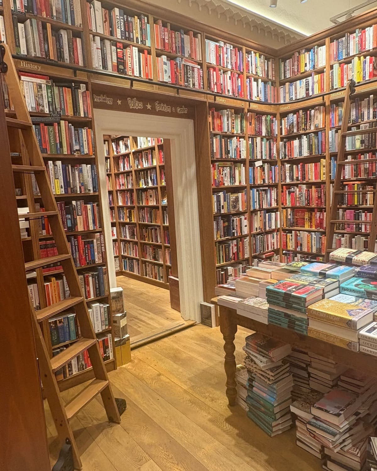 A bookshop room lined floor to ceiling, a ladder against the shelves and a table of stock in the middle.